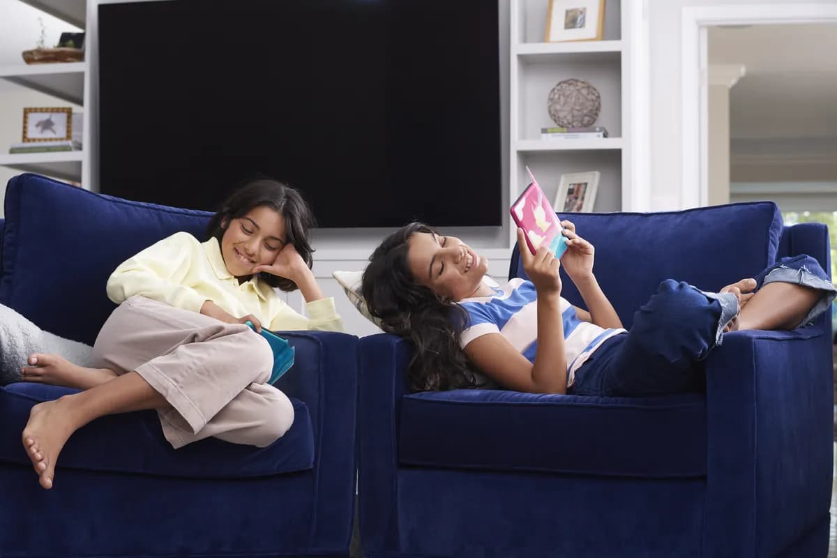 Two girls reading on a blue sofa, one holding a Kindle Kids in a patterned cover