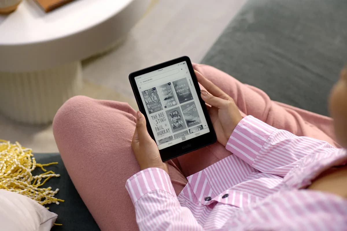 A reader on a sofa holding a Kindle Paperwhite with a page of book covers on screen