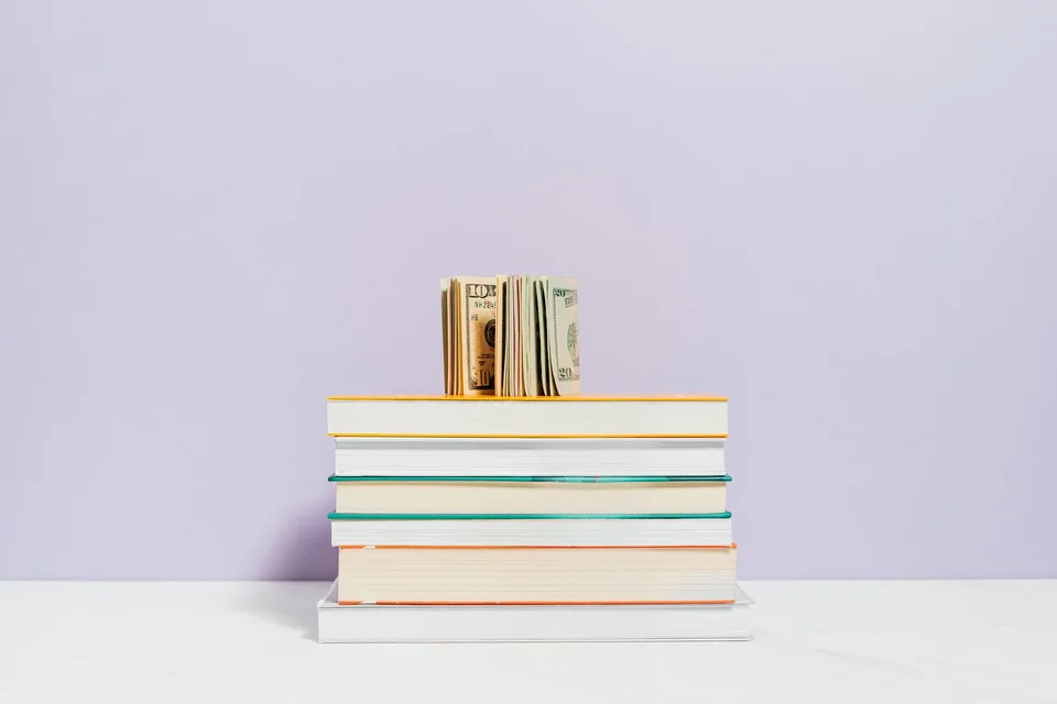 A stack of colorful books with folded dollar bills resting on top against a soft purple background