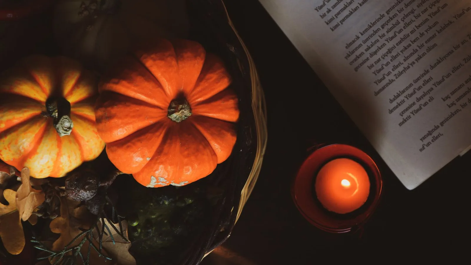 Carved pumpkins and a lit candle beside an open book on a dark table