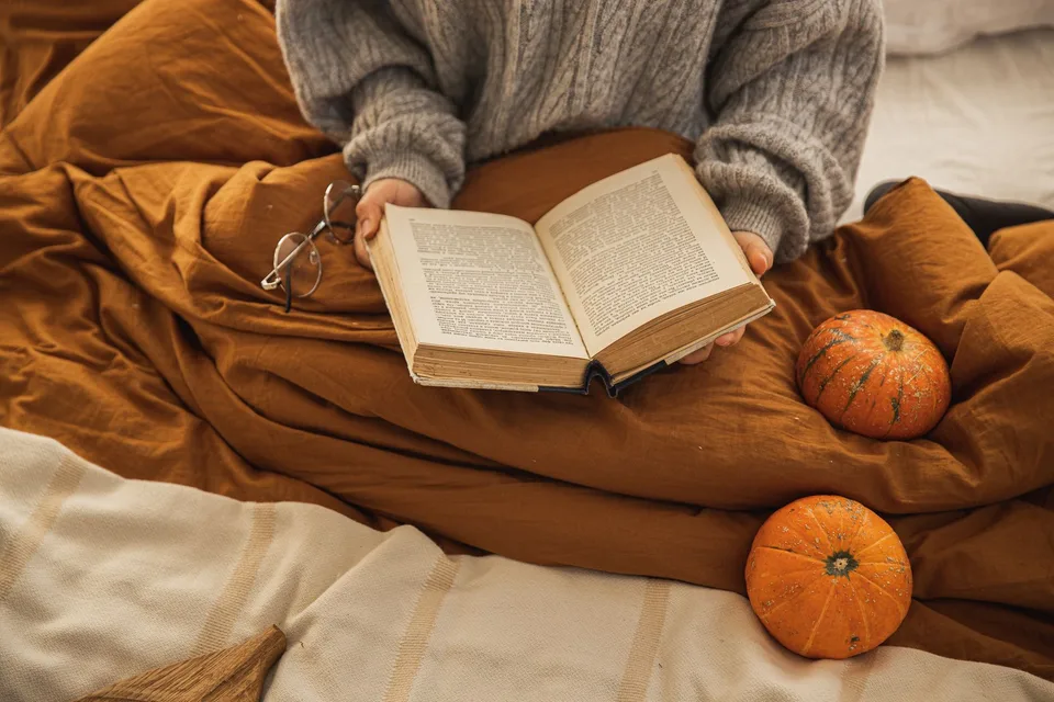 A person in a knit sweater reading a book in bed beside two small pumpkins