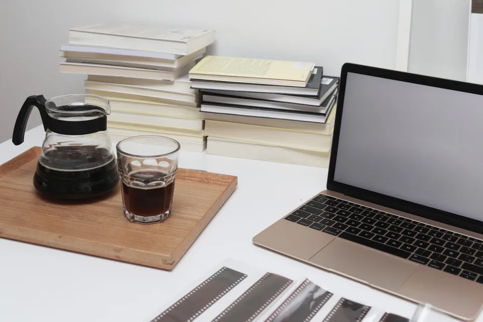 A laptop, stacked books, and coffee on a bright desk while preparing a book for publication
