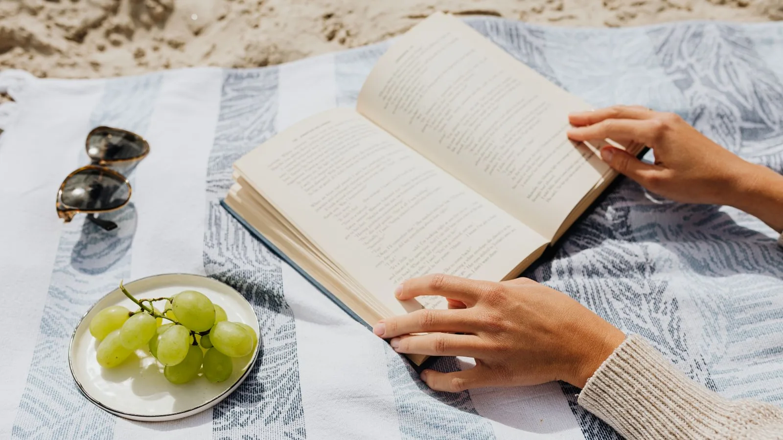 An open book on a striped beach towel beside sunglasses and a plate of grapes in bright summer sun